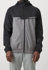 Svart och grå vindjacka med hel dragkedja framtill, elastiska ärmslut och huva; har reflexdetaljer på bröstet.