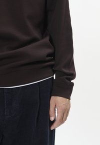 Brown cotton sweatshirt with a round neckline and ribbed cuffs, paired with dark corduroy pants. Hand positioned near the waist.