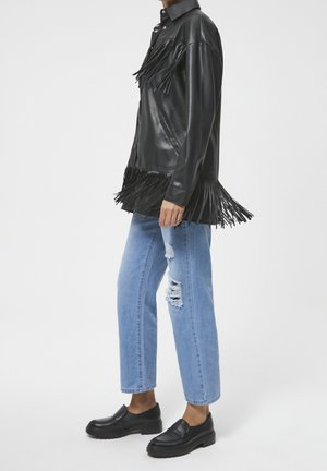 Veste en cuir noir avec détails à franges, associée à un jean bleu clair déchiré et des chaussures noires sans lacets. Texture lisse et design boutonné.