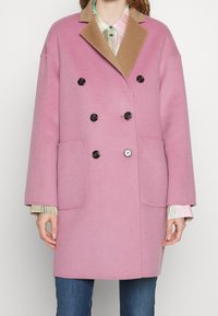 Dubbelzijdige jas in roze wol, met een tan kleurige kraag, zwarte knopen en zijzakken. Losse pasvorm met omhulde mouwen.