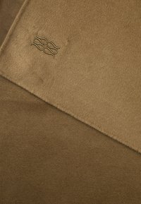 Nærbilde av kamelfarget ullstoff med brodert logo som har fire sammenkoblede sløyfer i matchende tråd.