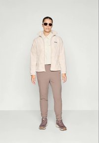 Chaqueta de forro polar en rosa pálido con cuello alto, combinada con una camiseta ligera y pantalones ajustados en color topo. Unos zapatos de senderismo marrones y gafas de sol completan el conjunto.
