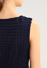 Námořnická modrá bezrukávová blůza s texturovanou tkaninou se vzorem ve tvaru diamantu. Design plátna zahrnuje na ramenou nařasené detaily pro zvýraznění.