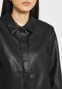 Giacca camicia in pelle nera con colletto a punta, chiusura con un bottone, superficie liscia e linee pulite. I dettagli sulla stoffa sono evidenti.