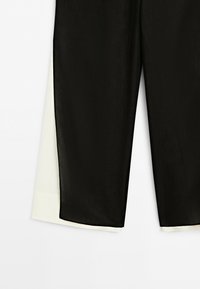 Close-up of black pants with a visible white fabric layer underneath at the hem, against a plain light background.