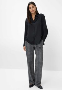 Camicia nera in satin con bottoni e maniche lunghe, abbinata a pantaloni grigi a quadri con gamba larga. L'outfit presenta una silhouette elegante e una texture morbida.