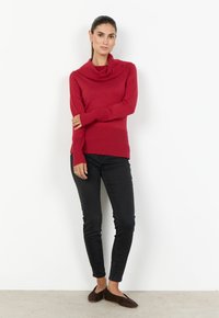 Kvinna står mot en vit vägg iförd en röd kofta med krage, svarta tight jeans och bruna lägenheter, med håret uppsatt och minimal smyckesstil.