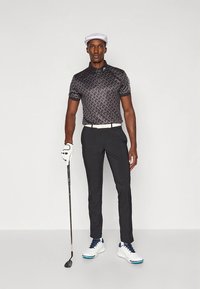 Črna polo majica z vzorci in kratkimi rokavi, kombinirana s črnimi hlačami po meri, belim pasom in golf čevlji. Drži golf palico in nosi belo kapo.