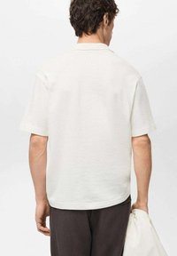 Polo bianco a maniche corte con tessuto strutturato. Presenta un colletto e un orlo leggermente curvo. Indossato con pantaloni scuri e tiene una borsa.