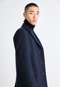 Marineblå ullfrakk med dobbelspent design, stor krage og fire knapper; brukt over en svart høyhalset genser. Jevn tekstur.