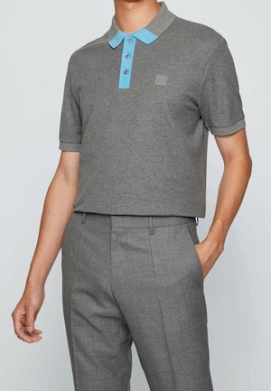 Mann trägt graues, kurzärmeliges Polo mit hellblauem Kragen und Knöpfen, kombiniert mit grauen, karierten, maßgeschneiderten Hosen.