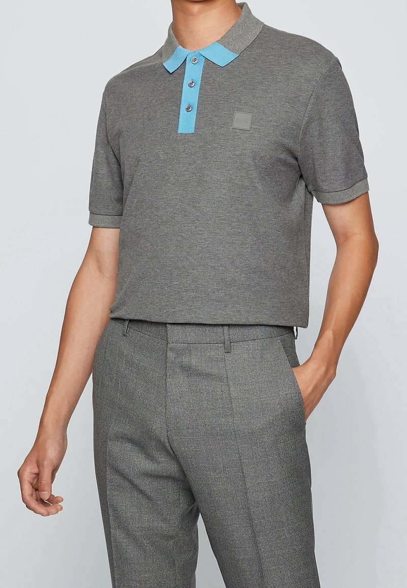 Homme portant un polo gris à manches courtes avec col et boutons bleu clair, assorti à un pantalon tailleur gris à carreaux.