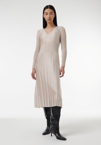 Robe longue plissée beige avec un décolleté en V et des manches longues. Elle présente de fines rayures verticales et est assortie à des bottes noires ajustées montantes jusqu'aux genoux.