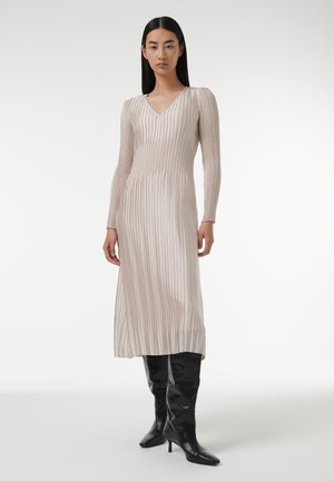 Robe longue plissée beige avec un décolleté en V et des manches longues. Elle présente de fines rayures verticales et est assortie à des bottes noires ajustées montantes jusqu'aux genoux.