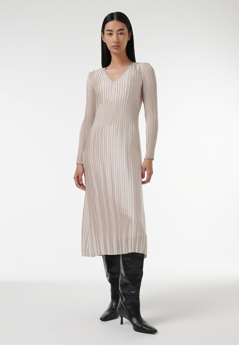 Robe longue plissée beige avec un décolleté en V et des manches longues. Elle présente de fines rayures verticales et est assortie à des bottes noires ajustées montantes jusqu'aux genoux.