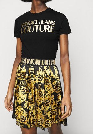 Žena nosící černé tričko Versace Jeans Couture a odpovídající černou a zlatou sukni s logem na pasu.