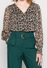 Blusa trasparente a maniche lunghe nera con stampa animale beige, abbinata a pantaloni verdi a vita alta con una cintura marrone circolare.
