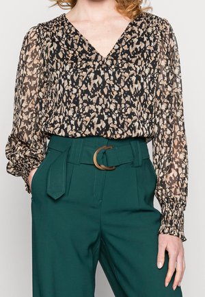 Blouse transparente à manches longues en noir avec un imprimé animal en beige, associée à un pantalon vert taille haute avec une ceinture ronde marron.