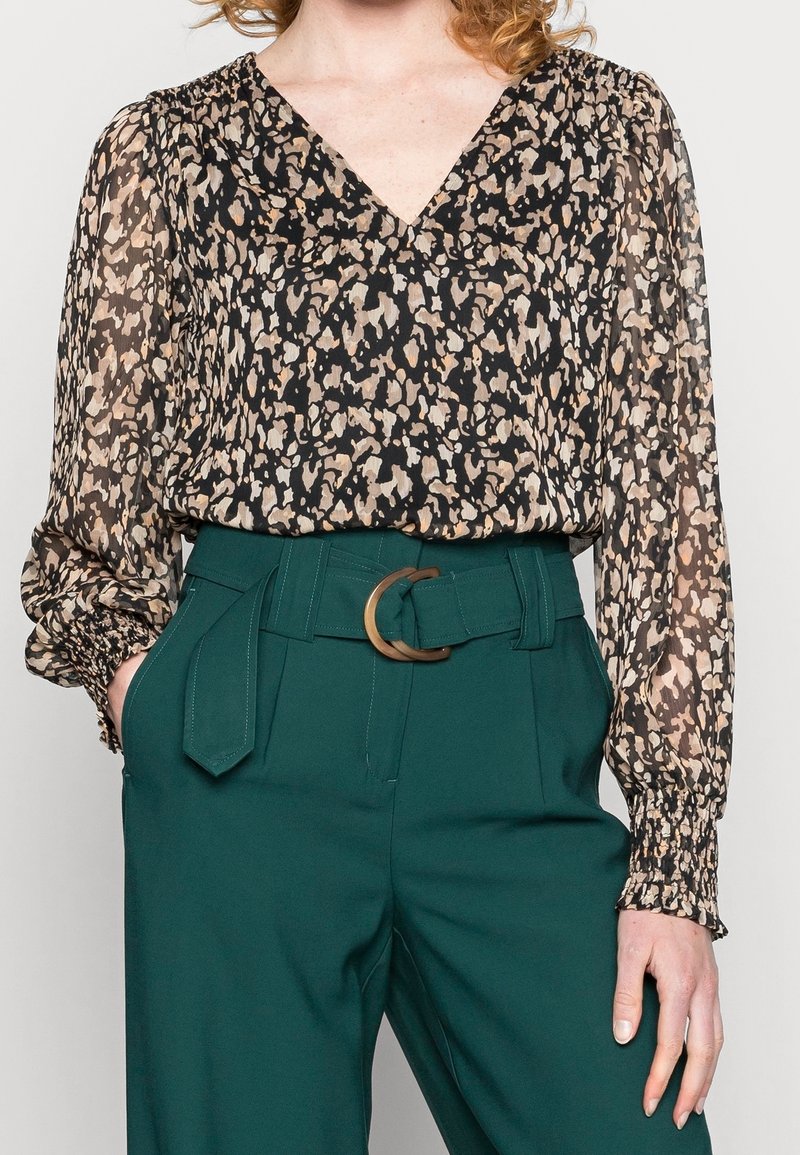 Blusa trasparente a maniche lunghe nera con stampa animale beige, abbinata a pantaloni verdi a vita alta con una cintura marrone circolare.