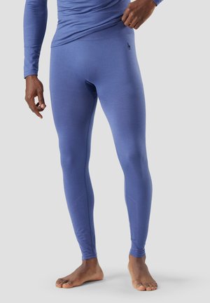 Man wearing fitted blue thermal leggings and long-sleeve top, adjusting waistband, standing barefoot on light gray background.