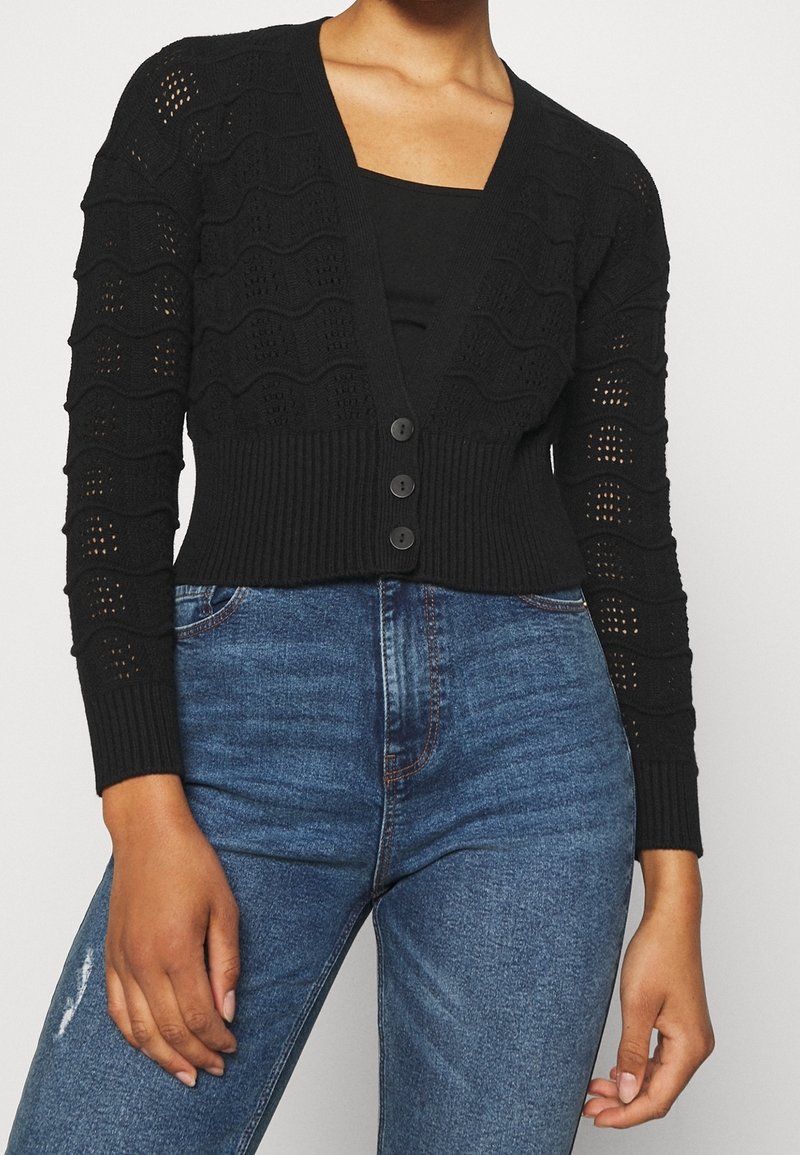 Femme portant un cardigan en maille texturée noire boutonné sur un haut noir avec un jean bleu taille haute.