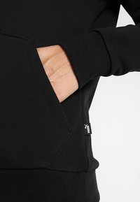 Sweat à capuche noir avec une poche kangourou. Présente des poignets et un bas côtelés, une texture douce en mélange de coton, avec une petite étiquette logo Puma sur le côté.