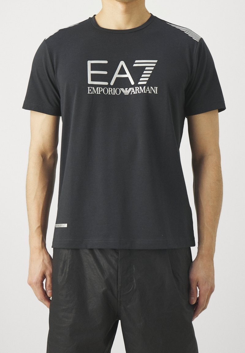 Musta puuvillainen t-paita lyhyillä hihoilla, jossa on hopeinen "EA7 EMPORIO ARMANI" -logo. Sisältää raitakoristeet hartioilla.