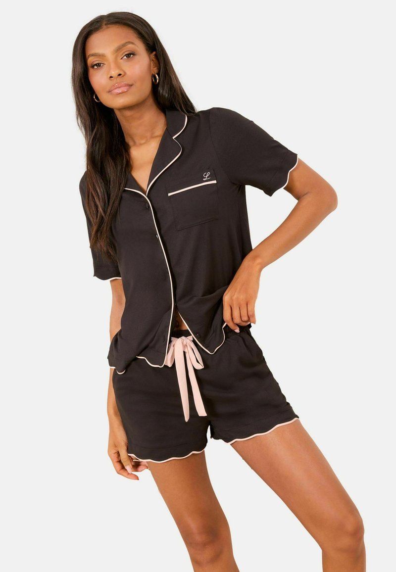 Femeie purtând pijama neagră cu mâneci scurte și pantaloni scurți, cu tiv roz pal și șnur, pozând cu o mână pe șold.