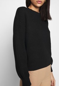 Schwarzer, gerippter Strickpullover mit rundem Halsausschnitt und langen Ärmeln, mit einem enganliegenden Saum. Die Textur des Pullovers ist erhaben, der Stoff wirkt dick und warm.