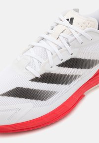 Valkoinen urheilukenkä, jossa on verkkopäällinen, mustat korostaraidat ja kirkkaanpunainen kumipohja. Kenkässä on nauhat ja brändäys kielen kohdalla.