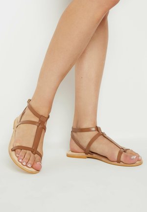 Bruine leren sandalen hebben een T-bandsluiting, een platte zool en verstelbare enkelbandjes. De textuur is glad met een minimalistische uitstraling.