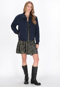 Sötétkék farmerdzseki gombos elülső résszel, bordázott mandzsettákkal és két oldalsó zsebbel, virágmintás ruhára és fekete térd fölé érő csizmára rétegezve.