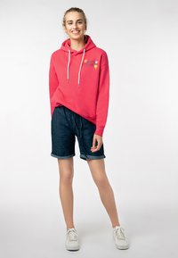 Roze hoodie met trekkoorden en florale borduursels, gecombineerd met donkere denim shorts. Model draagt witte sneakers, wat een casual outfit benadrukt.