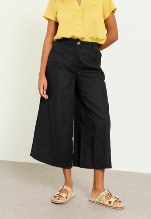 Frau trägt schwarze, weit geschnittene, kurze Hosen, gelbe Kurzarmbluse und beigefarbene Sandalen mit bunten Fruchtmustern.