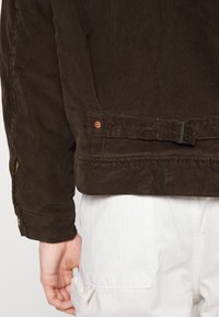 Veste en velours côtelé marron foncé avec sangle latérale réglable et rivet en cuivre, portée sur un pantalon blanc avec poche arrière et détail boucle.