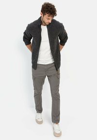 Dunkelgrauer Reißverschluss-Pullover mit Rippkragen, kombiniert mit hellgrauen Cargo-Hosen mit mehreren Taschen und hellbraunen Akzenten. Darunter ein weißes T-Shirt.