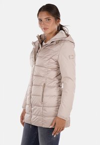 Giacca puffer beige con cappuccio, texture trapuntata e chiusura frontale a zip. Presenta tasche laterali e un patch con logo sulla parte superiore del braccio.