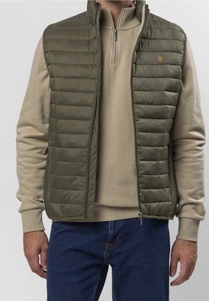 Mand iført olivengrøn quiltet vest over beige zip-up sweater og blå jeans mod en ensfarvet lys baggrund.