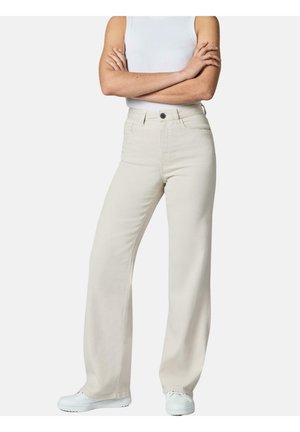 Person, der står med armene over kors iført højtaljede beige bredbenede bukser, hvid tanktop og hvide sneakers mod en ensfarvet baggrund.