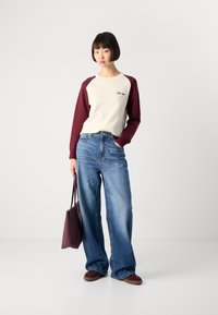 Woman in cream and burgundy long-sleeve top, wide-leg blue jeans, burgundy shoes, and holding a burgundy tote bag, standing against a white background.