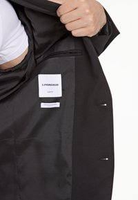 Schwarzer Blazer mit einem glatten Satinfutter, ausgestattet mit einem Slim-Fit-Etikett, einer Innentasche und einem eleganten Design. Minimalistische Hardware-Akzente sind vorhanden.