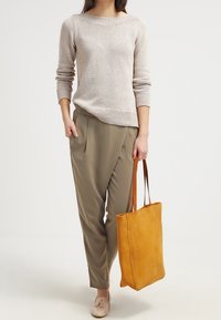 Beiger Strickpullover mit weitem Halsausschnitt, olivgrüne schmal zulaufende Hose, beige Slipper und eine strukturierte gelbe Tote-Tasche mit braunen Griffen.