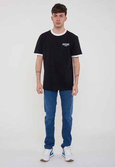 Maglietta nera con colletto e bordi delle maniche bianchi, logo sul petto, abbinata a jeans blu e sneakers bianche con accenti blu.