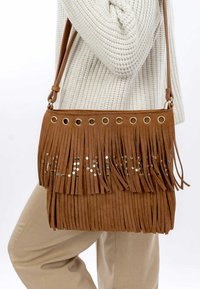Bolso bandolera de ante marrón con detalles de flecos, tachuelas plateadas y ojales dorados. Correa ajustable y textura suave.