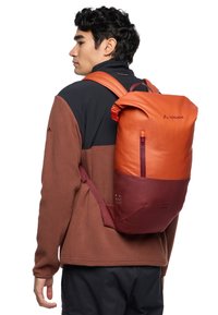 Mochila naranja y marrón con diseño de parte superior enrollable, correas ajustables y bolsillo frontal con cremallera. Fabricada con material texturizado y resistente a las inclemencias del tiempo.
