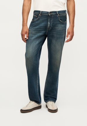 Homme portant un jean droit bleu délavé et des baskets blanches, debout devant un fond clair uni.