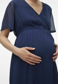 Tamnoplava haljina za trudnice s kratkim, lepršavim rukavima i dizajnom koji se obavija. Mekani, teksturirani materijal pada preko zaobljenog trbuha. Ruke nježno odmaraju.