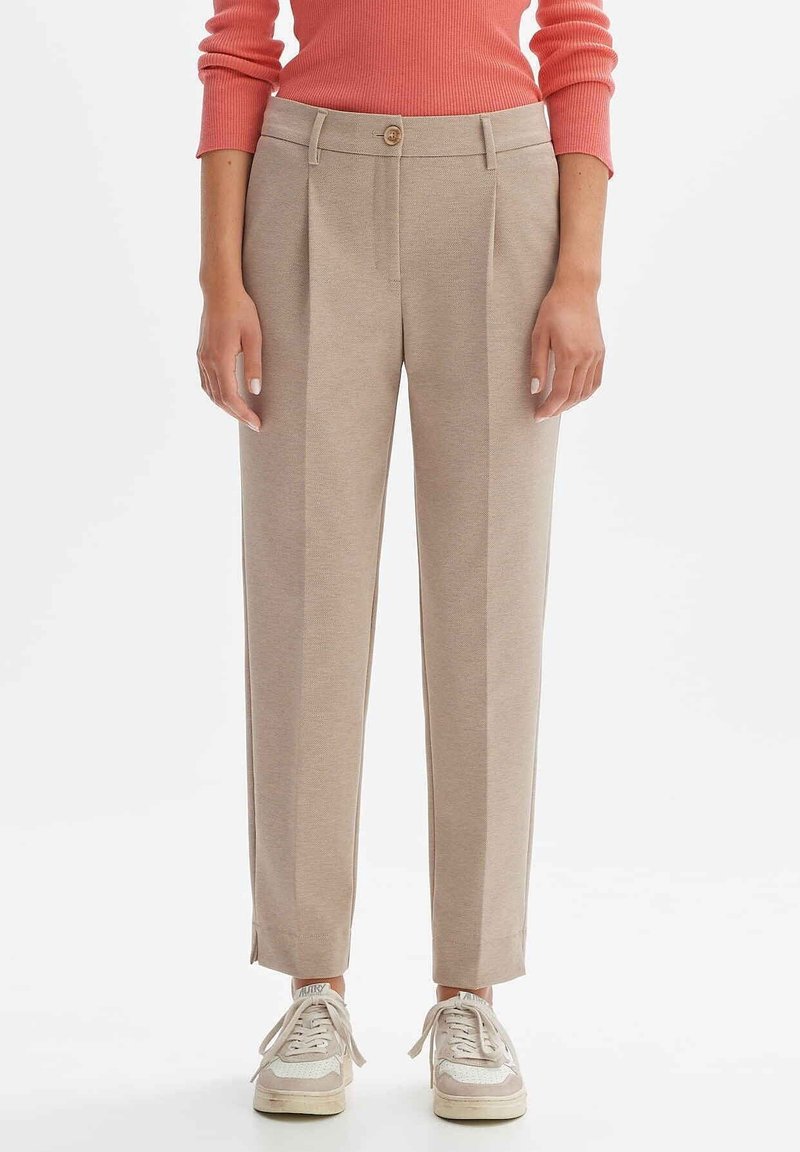 Femme portant un pantalon beige droit avec des plis devant, fermeture à boutons, associé à des baskets blanches et un pull côtelé corail.