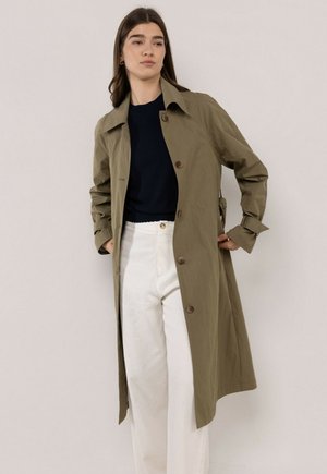 Mujer con un abrigo trench verde oliva abotonado, top negro y pantalones blancos de cintura alta, de pie frente a un fondo claro y liso.