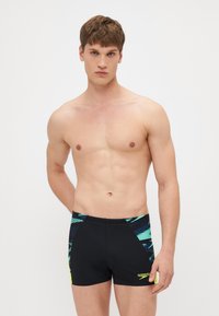 Crne kupaće gaće s tirkiznim i plavim geometricnim uzorcima, kontrastni žuti logo, elastična waistband i prilagođeni dizajn.